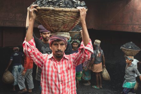 Worker in a light red checkered shirt holding onto a basket of coal on the top of their head with two hands Heavy baskets of coal are transported on the workers' heads in hot conditions at Dhaka, Bangladesh.