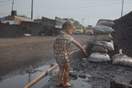 A child in a checkered shirt plays on a wet gravel road while the parents work in the coal mine A Dhaka, Bangladesh coal worker child finds something of interest near the huge coal pile.
