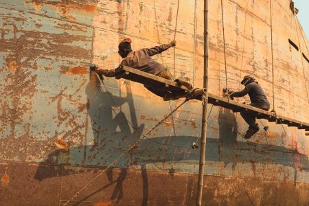 The workers are held in the air precariously by ropes and a wooden platform to access the vessel's side body. Therefore, they can sand and grind the vessel. Hanging precariously off platform during repairs at Dhaka, Bangladesh Shipyard.