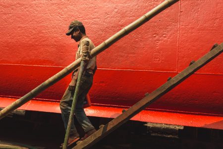 Shipyard by the river Buriganga in Dhaka, Bangladesh. A man walking down on a wooden bamboo ladder walks past a vibrant red wall, his silhouette beautifully outlined against the bold colour. The composition captures a moment of quiet determination, blending the rich texture of the wall with the subject's gritty reality, resilience, and hard work.