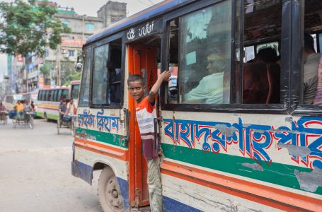 Clinging to a battered bus, a young boy calls for passengers. This is not a childhood filled with play—it’s a shift that runs from dawn to dusk. A young boy in Dhaka, Bangladesh, clings to the open door of a worn-out public bus, his face showing signs of exhaustion and determination. He works as a bus helper, a common form of child labour in the city. The faded paint and peeling letters on the bus reflect the harsh urban environment and the chaotic streets in the background highlighting the struggles of daily life.