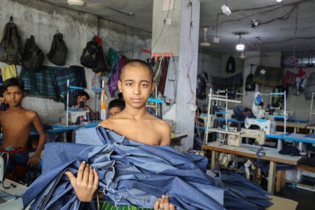 In a cramped garment factory in Dhaka, a young boy stands shirtless, arms full of denim. Around him, other boys stitch quietly, invisible threads in the global supply chain. A shirtless boy holds a large bundle of denim fabric inside a crowded garment factory in Bangladesh, surrounded by other child workers and sewing machines. His gaze is direct, exposing the difficult conditions and exploitation faced by children in the clothing industry.