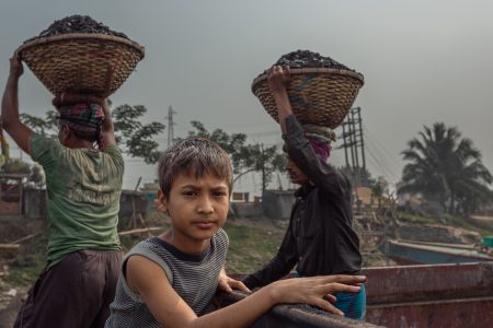 A boy leans on a container in a coal yard, dwarfed by labourers and their load. A young boy in a striped sleeveless shirt leans on the edge of a rusty container, gazing into the camera at a coal industry site in Bangladesh. Behind him, two labourers carry large woven baskets filled with coal on their heads, walking through the dusty industrial landscape. Power lines, trees, and a hazy sky frame the background, emphasizing the harsh working conditions