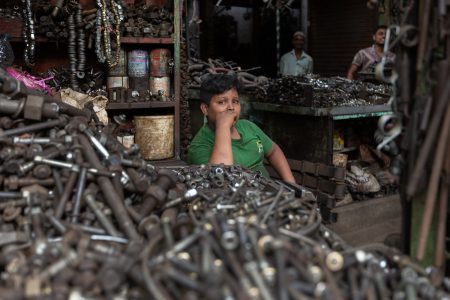 Behind pipes and bolts, a boy in green pauses—his burden heavier than the scrap around him. A young boy in a green t-shirt sits behind a pile of metal bolts and spare parts in a busy workshop in Bangladesh. His hand rests on his face as he looks into the distance, surrounded by industrial tools and machinery. The background shows adult workers and shelves stacked with mechanical parts. As a child labourer, he spends his days selling spare parts instead of attending school, highlighting the ongoing issue of child labour in hazardous work environments.
