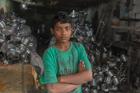 A boy stands among aluminium pots—sweat and grime revealing untold labour stories. A young boy stands in front of an aluminium pot-making factory in Bangladesh, his arms crossed and his expression serious as he looks directly into the camera. Wearing a worn green t-shirt, he appears strong and tired, a sign of the harsh labour conditions he endures. Behind him, the industrial setting is filled with metal pots and machinery, emphasizing the harsh environment where child labour persists.