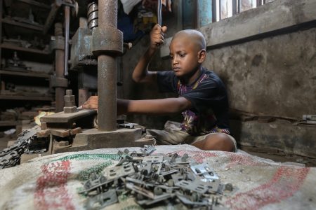 In a dim metal workshop, a boy operates a heavy press. The jagged parts he produces are for locks and hinges, but their cost is unseen. A young boy with a shaved head and a serious, focused expression operates a heavy metal press inside a dimly lit factory in Bangladesh. He wears a black and purple patterned t-shirt and beige shorts, sitting on the ground as he uses both hands to manipulate the machine. In the foreground, a pile of metal components rests on a worn-out sack, adding to the industrial setting. The background reveals rusted machinery, tools, and a weathered concrete wall with a barred window, allowing a small amount of natural light to filter in. This image highlights the harsh realities of child labour in dangerous working conditions.