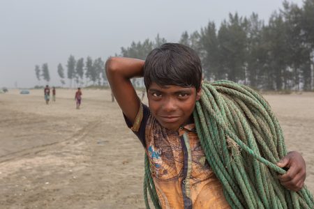 A young boy with dark, damp hair and a dirt-streaked face carries a heavy, sand-covered green rope over his shoulder while working with fishermen in Cox’s Bazar, Bangladesh. A young boy with dark, damp hair and a dirt-streaked face carries a heavy, sand-covered green rope over his shoulder while working with fishermen in Cox’s Bazar, Bangladesh. His worn and stained shirt features a faded world map design. He gazes directly into the camera with a determined yet subtle smile. In the background, a hazy shoreline with scattered trees and distant figures highlights the vastness of his workplace—the beach where labour replaces childhood