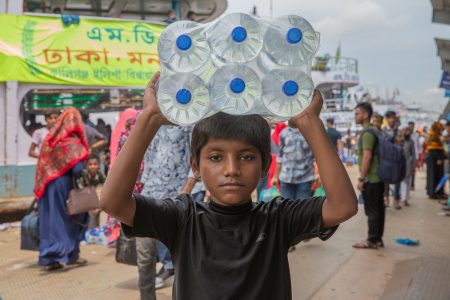 At Sadarghat port, a boy weaves through crowds with water bottles—survival starts early here. A young boy in Bangladesh carries a heavy pack of bottled water on his head while standing at a busy port. Dressed in a black t-shirt, he looks directly into the camera with a serious expression, reflecting the hardships of child labour. Behind him, a crowd of travellers moves in different directions, highlighting the bustling environment where children like him work to make a living