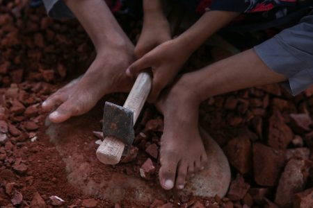 A child breaks bricks into rubble with a hammer, bare feet in red dust. These fragments, sold for construction, are built on shattered youth. A young girl works barefoot in a brick field in Bangladesh, using her feet to stabilize a large piece of brick while she grips a worn hammer with both hands. She carefully strikes the brick to break it into smaller pieces, highlighting the harsh realities of child labour in the brick-making industry. The rough, red clay dust covers the ground beneath her, emphasizing the physical toll of her work.
