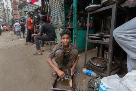 In a mechanic’s alley, a barefoot boy cleans metal parts—one of many who work to survive instead of study. A young boy squats on the side of a busy street in Bangladesh, scrubbing metal parts in a small tray of murky water. Dressed in worn-out clothes, he looks directly at the camera with a quiet resilience. Behind him, other workers and pedestrians move through the bustling environment of an auto parts market. The image captures the harsh reality of child labour, where children work instead of attending school, highlighting the ongoing struggle for their rights and well-being