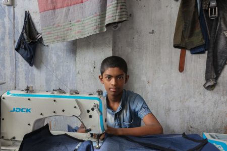 Inside a narrow garment workshop, a boy concentrates on a sewing machine, stitching denim with the precision of a seasoned worker. A young boy in a denim shirt sits in front of an industrial sewing machine, working on fabric inside a garment workshop in Bangladesh. His serious expression and the worn-out surroundings highlight the harsh reality of child labour in the textile industry.