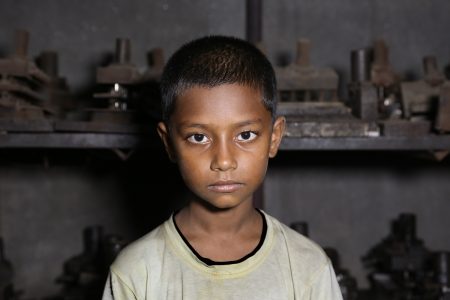 A boy faces the camera before heavy machinery, silent yet powerful—an emblem of Bangladesh's hidden child labor. A young boy with short black hair and deep brown eyes stands before industrial metal machinery in a dimly lit factory in Bangladesh. His expression is serious and sombre, and his face is illuminated by a direct light source, highlighting the contrast between his youthful features and the harsh working environment. He wears a worn-out pale yellow T-shirt with a black collar, slightly stained from labour. The background consists of heavy, rusted metal equipment, indicating the setting of a metal factory.