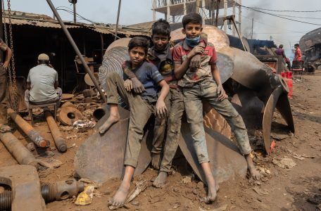 Three boys, faces and shirts blackened by grit, sprawl atop a massive metal propeller in a foundry. Their eyes, ringed with tiredness and longing, linger on a rare moment of rest amid the furnace heat. Three young boys covered in dust and dirt sit on a massive metal ship propeller at a foundry in Bangladesh. Their faces and clothes are smeared with grime, and they are barefoot, resting on the industrial scrap. One boy wears a tattered blue and red shirt, another a checkered shirt with a British flag emblem, and the third a dark, stained shirt. One has a face mask pulled down around his neck. They stare directly at the camera with expressions that blend resilience, exhaustion, and camaraderie. Behind them, workers labour amidst heavy machinery, rusted metal, and smoke rising from the factory, highlighting the harsh reality of child labour in the foundry industry