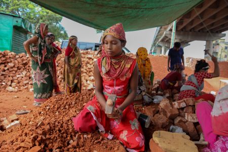 A girl in vivid red hammers bricks atop a mound, surrounded by peers under relentless heat. A girl wearing a red dress and a patterned scarf sits on a mound of broken bricks at a brickfield in Bangladesh. She holds a hammer in her hand, looking downward solemnly. Behind her, other women and children work under a makeshift tarp, surrounded by bricks and dust.