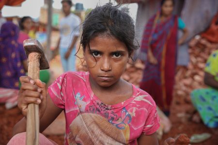 Her eyes meet the camera with unflinching intensity. Dressed in a bright pink shirt, she grips a hammer used to crush bricks . A young girl with dust-covered skin and intense eyes grips a worn hammer while working in a brick field in Bangladesh. She wears a faded pink shirt with a cartoon print, contrasting with the harsh labour she endures. Behind her, women and other workers move among piles of broken bricks. Her determined, solemn expression reflects the harsh reality of child labour in the brick-making industry.